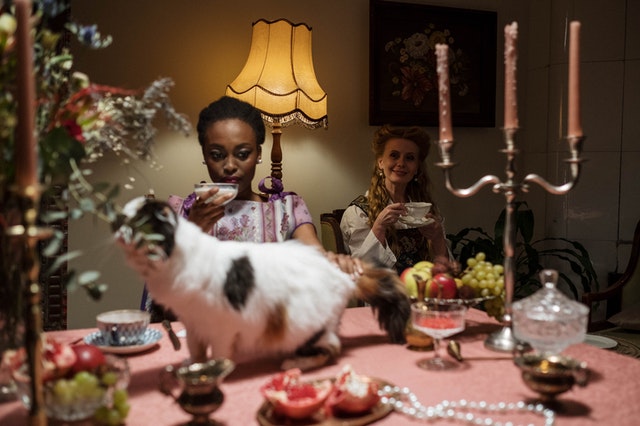 Two women sitting at a table with fruits, a cat, and a candle holder on it.