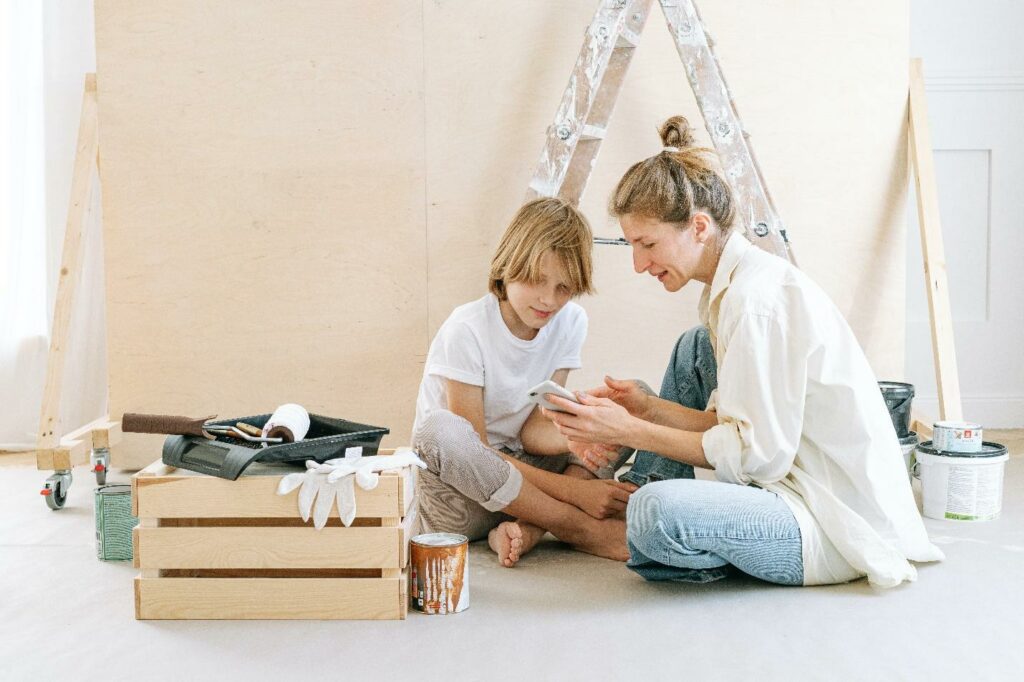 woman and preteen look at phone next to cans of paint, ladder, and construction materials